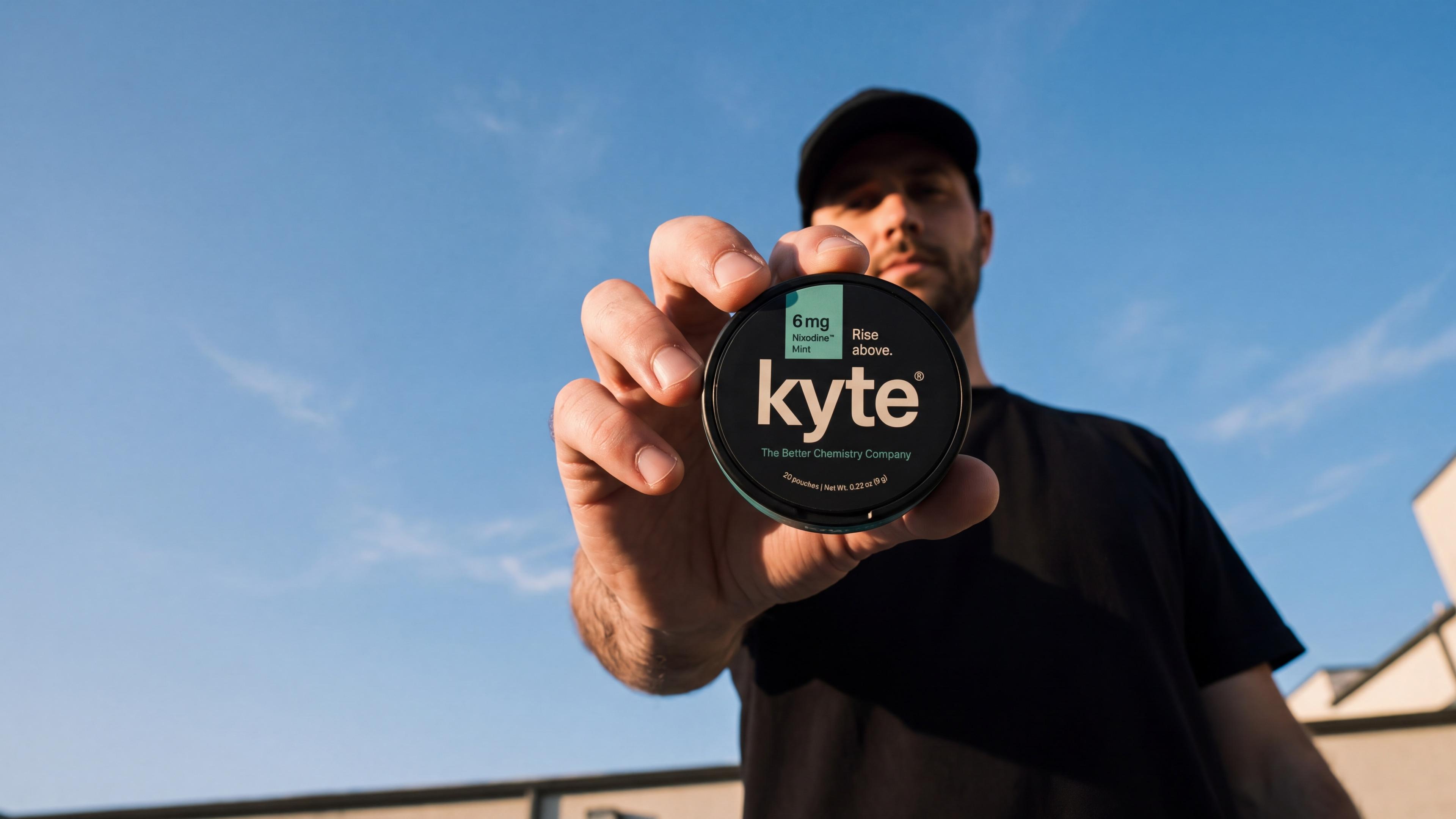 Man holding a Kyte Mint tin toward the camera against a blue sky